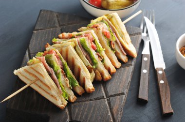 Tahta bir tahtanın üzerinde beyaz ekmekli bir sandviç. Sandviç dört bölüme ayrılır ve ahşap bir şiş üzerine sabitlenir. Yakınlarda çatal bıçak takımı var. Koyu ahşap arka plan. Close.