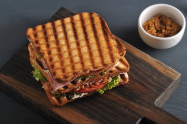 Tahta bir tahta üzerinde beyaz ekmek birkaç dilim Sandviç. Sandviçin dolgusu pastırma, peynir, jambon, sos, domates ve maruldan oluşur. Hardallı bir sonraki bardak. Karanlık arka plan. Close.