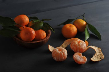 Ahşap bir kapta yeşil yaprakları ile Mandalina. Deride kabuksuz mandalina ve mandalinanın yarısı vardır. Koyu ahşap arka plan. Yukarıdan görüntüleyin. Close.