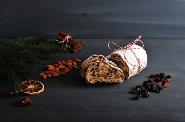 Christmas shtollen cut into slices. The composition is complemented by a spruce branch, various spices, nuts, raisins, and a dry citrus slice. Dark wooden background. Close-up. clipart