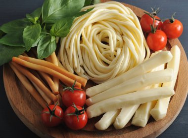 Peynir tahta bir tabakta sürünüyordu. Peynir spagetti şeklinde, pipet şeklinde füme ve beyaz. Çeri domates, fesleğen yeşillikleri yanında. Closeup. Yukarıdan görüntüleyin. Makro fotoğrafçılık.