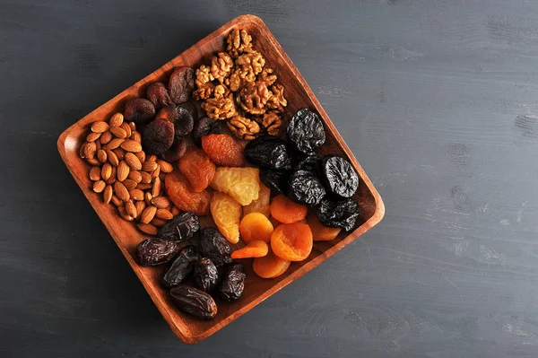 Kurutulmuş meyve ve fındık bir tabakta. Tabakta hurma, kuru erik, kuru kayısı, kuru armut ve şeftali, badem ve ceviz bulunmaktadır. Karanlık bir arka plan. Yukarıdan görüntüleyin. Close. Makro fotoğrafçılık.