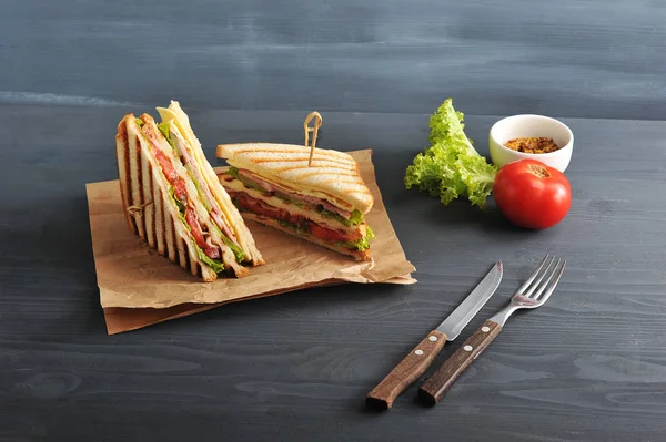 Two halves of a sandwich on kraft paper. The stuffing of the sandwich club consists of ham, cheese, tomato, bacon, sauce and fried egg. Nearby cutlery, mustard, vegetables. Dark background. Close-up.