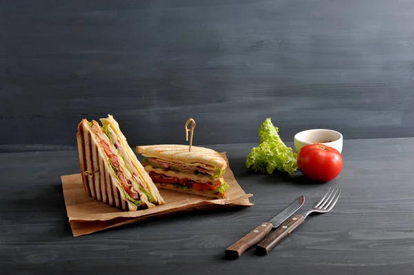 Two halves of the club sandwich on kraft paper. The filling of the sandwich consists of ham, cheese, tomato, bacon, sauce and fried egg. Nearby cutlery, mustard, vegetables. Dark background. Close-up.