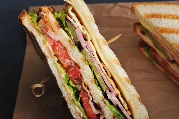 Club sandwich on kraft paper. On the cut of the sandwich, the filling of tomato, ham, cheese, bacon, lettuce, cucumber and fried egg is clearly visible. Dark background. Close-up. Macro photography.