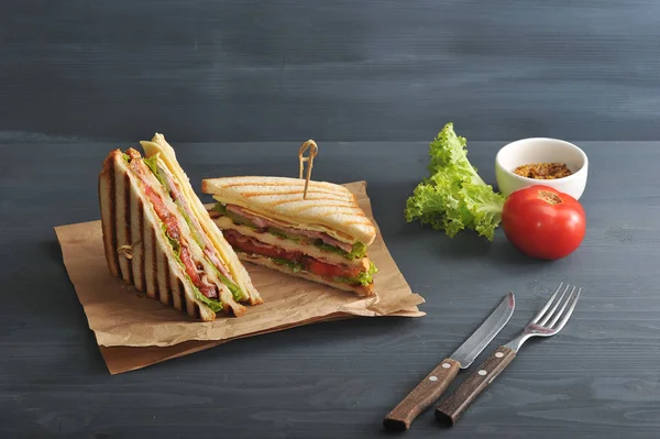 Two halves of a sandwich on kraft paper. The stuffing of the sandwich club consists of ham, cheese, tomato, bacon, sauce and fried egg. Nearby cutlery, mustard, vegetables. Dark background. Cl