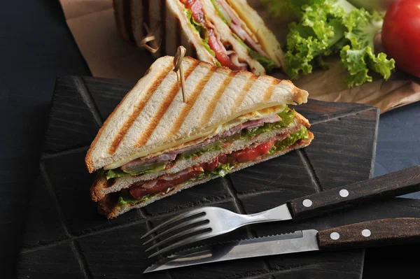 Half a club sandwich on a wooden board. The other half of the sandwich is next to kraft paper. In the frame cutlery, mustard, lettuce, tomato. Dark background. Close-up. Macro photography.