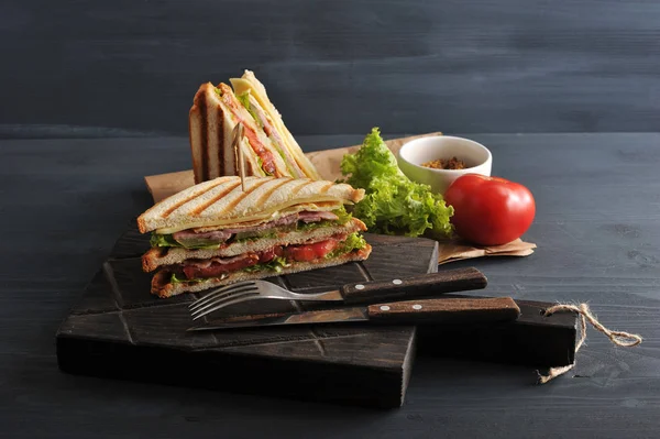 Half a club sandwich on a wooden board. The other half of the sandwich is next to kraft paper. In the frame cutlery, mustard, lettuce, tomato. Dark background. Close-up.