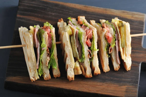 Original sandwich feed. On a wooden board, a sandwich is cut into four pieces diagonally. Pieces are fixed on a wooden skewer. Dark wooden  background. Close-up. Macro photography.