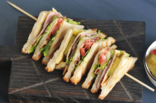 Sandviç dört bölüme ayrılır ve ahşap bir şiş üzerine sabitlenir. Sandviçin dolgusu pastırma, peynir ve jambondan oluşur. Koyu ahşap arka plan. Close.