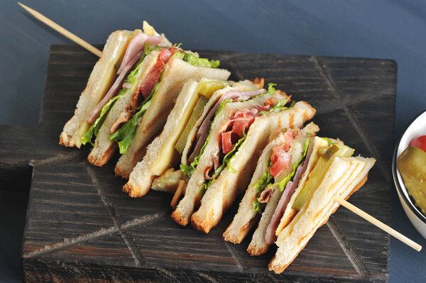 Сэндвич разделен на четыре части и закреплен на деревянном шампуре. Наполнитель сэндвича состоит из бекона, сыра и ветчины. Темный деревянный фон. Крупный план
.