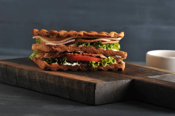Sandwich of white bread on a wooden board. The filling of the sandwich consists of bacon, cheese, ham, sauce, tomato and lettuce. Dark wooden background. Close-up. Macro photography.