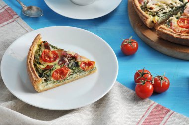Ispanak, pastırma, çeri domates ve peynirile pasta quiche bir parça. Çerçeveiçinde, bir tabak ve kiraz domates üzerinde kek bir parça. Mavi arka plan bir peçete ile kaplı. Close.