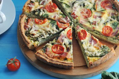 Ahşap bir tabak, ıspanak ve pastırma ile pasta quiche. Yemek pişirirken peynir ve domates kullanılır. Çerçevede, bir kiraz domates. Mavi arka plan. Yukarıdan görüntüleyin. Close. Makro fotoğrafçılık.