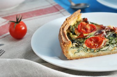 Beyaz bir tabakta Fransız pasta quiche bir kısmı. Ispanak, pastırma, peynir, kiraz domatesi ile dolum. Çerçevede, bir kiraz domates. Arka plan bir peçete ile kaplıdır. Close. Makro fotoğrafçılık.