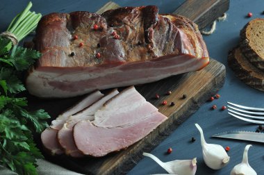 Tahta bir tahtanın üzerine bir parça pastırma ve birkaç doğranmış dilim. Çerçevede, maydanoz, sarımsak, ekmek ve çatal bıçak takımı bir demet. Koyu arka plan peçete ile dekore edilmiştir. Close. Makro fotoğrafçılık.