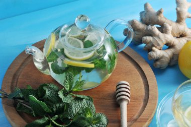 Cam bir çaydanlıkta zencefil içeceği. Nane, zencefil, ballı limondan yapılmış faydalı bir içecek. Çerçevede zencefil ve nane bir demet kökü. Mavi ahşap arka plan. Close.
