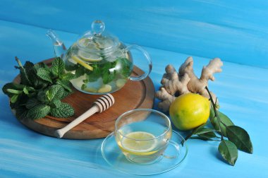 Cam bir çaydanlıkta zencefil içeceği. Bir sonraki bardak ve bir içki. Çerçevede, zencefilkökü, bir demet nane, limon. Mavi ahşap arka plan. Close.