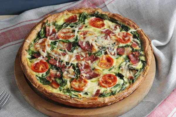 Tahta bir tabakta, pastırma ve ıspanak ile bir pasta quiche. Yemek pişirirken, peynir ve domates dekorasyon için kullanılır. Arka plan bir bez peçete ile kaplıdır. Close. Makro fotoğrafçılık.