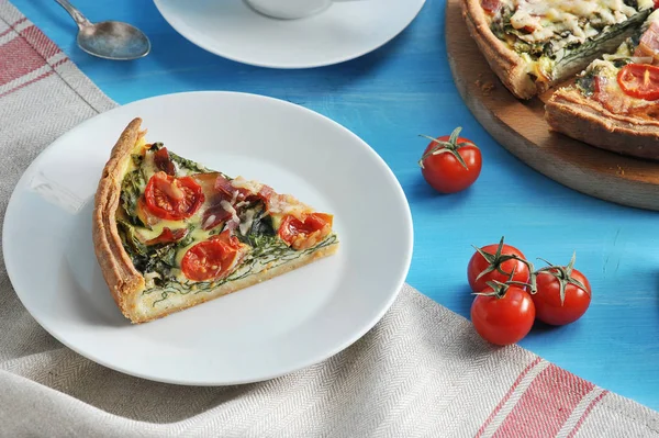 Ispanak, pastırma, çeri domates ve peynirile pasta quiche bir parça. Çerçeveiçinde, bir tabak ve kiraz domates üzerinde kek bir parça. Mavi arka plan bir peçete ile kaplı. Close.