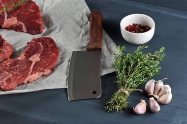 Parşömen kağıdında çiğ biftek. Et kesmek için biftek bıçağının yanında. Çerçevede, bir demet kekik, sarımsak, bir fincan biber. Koyu ahşap arka plan. Close.