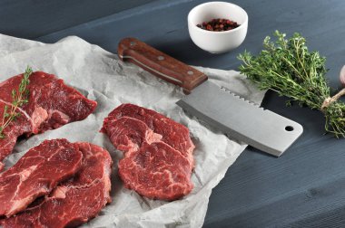 Kağıt üzerinde çiğ biftek. Bifteklerin yanında et için bir kesme bıçağı var. Çerçevede, bir demet kekik ve bir fincan biber. Koyu ahşap arka plan. Close. 