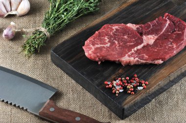 Tahta bir tahtada çiğ biftek. Biftek yanında biber ve büyük tuz vardır. Çerçevede, kekik, sarımsak ve et için bir kesme bıçağı bir demet. Pürüzlü bir tuvalin yüzeyi. Makro fotoğrafçılık.