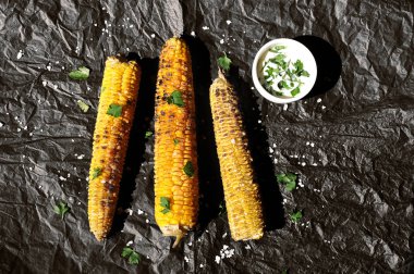 Izgarada pişirilmiş üç mısır koçan. Mısır otlar ve büyük deniz tuzu serpilir. Beyaz soslu bir sonraki fincan. Siyah arka plan. Yukarıdan görüntüleyin. Close.