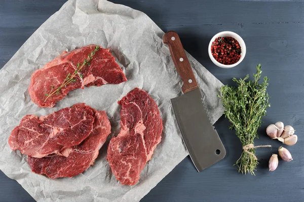 Raw beef steaks on paper. On one of the steaks is a twig of thyme. In the frame, a bunch of thyme, a garlic head, a cup of pepper and a cutting knife for meat. Dark background. View from above.