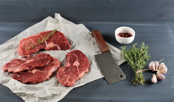 Kağıt üzerinde çiğ biftek. Bifteklerden birinde bir dal kekik var. Çerçevede, bir demet kekik, bir sarımsak kafası, bir fincan biber ve et için bir kesme bıçağı. Koyu arka plan. 