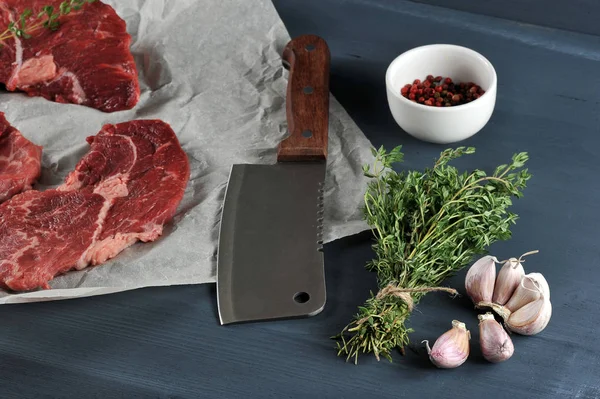 Parşömen kağıdında çiğ biftek. Et kesmek için biftek bıçağının yanında. Çerçevede, bir demet kekik, sarımsak, bir fincan biber. Koyu ahşap arka plan. Close.