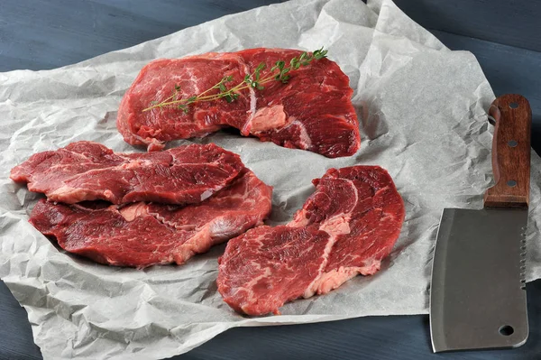 Raw beef steaks on paper. On one of the steaks is a twig of thyme. Next to the steaks is a cutting knife for meat. Dark wooden background. Close-up.