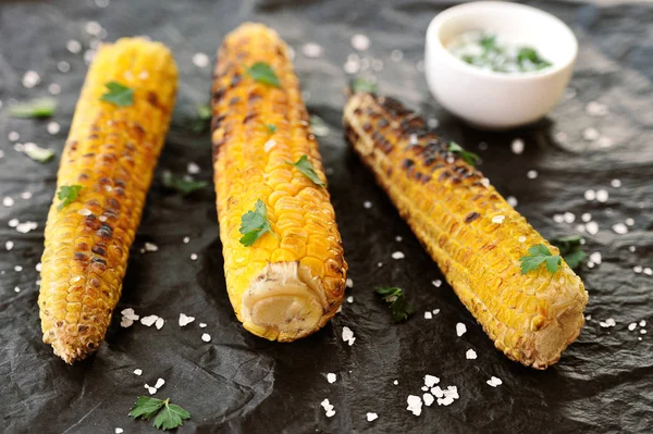 Izgarada pişirilmiş üç mısır koçan. Mısır otlar ve büyük deniz tuzu serpilir. Beyaz soslu bir sonraki fincan. Siyah arka plan. Close.