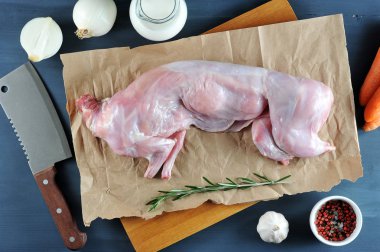 Biberiye ile tavşan pişirmek için bir set. Çerçevede, kraft kağıt üzerinde ham bir tavşan. Yakındaki havuç, soğan, biberiye, sarımsak, sürahi krema, bıçak. Karanlık arka plan. Yukarıdan görüntüleyin. Close.