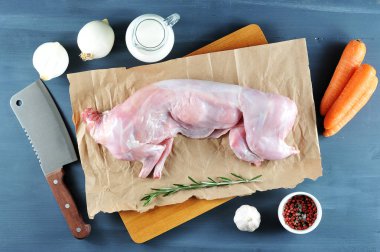 Biberiye ile tavşan pişirmek için bir set. Çerçevede, kraft kağıt üzerinde ham bir tavşan. Yakındaki havuç, soğan, biberiye, sarımsak, kremalı sürahi, et kesmek için bıçak. Karanlık arka plan. Yukarıdan görüntüle. 