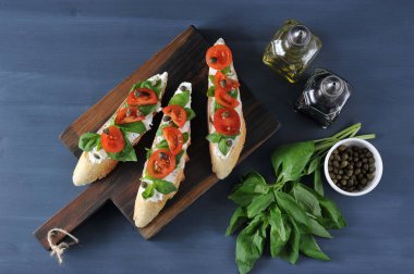 Bruschetta yumuşak peynir, taze domates, fesleğen yaprakları ve kapari ahşap bir tahta üzerinde. Çerçeveiçinde, zeytinyağı, balzamik sirke, fesleğen ve kapari ile bir kap. Karanlık arka plan. Yukarıdan görüntüle.