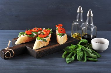Bruschetta yumuşak peynir, taze domates, fesleğen yaprakları ve kapari ile. Geleneksel İtalyan atıştırmalıkları. Çerçeveiçinde, zeytinyağı, balzamik sirke, fesleğen ve kapari ile bir kap. Koyu arka plan.