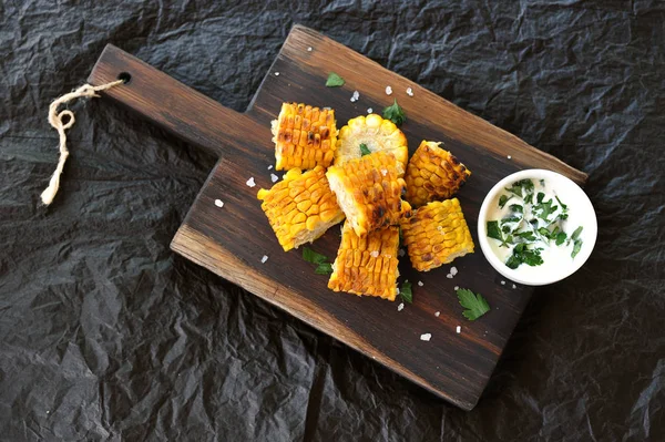 Izgarada pişirilmiş mısır. Ahşap bir tahta üzerinde mısır koçan dilimleri otlar ve deniz tuzu serpilir. Bir sonraki soslu fincan. Siyah arka plan. Close. Yukarıdan görüntüle.