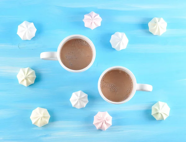 Two cups of hot cocoa. A fragrant drink energizes and creates a mood. Round the cups are arranged meringues. Blue wood background. View from above. Close-up.