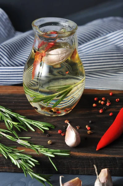 Tahtada zeytinyağı olan bir sürahi var. Yağ da biberiye, biberiye ve sarımsak yüzer. Sonra biberiye, sarımsak karanfil, acı kırmızı biber bir dal. Arka planda bir peçete. Dikey çekim.