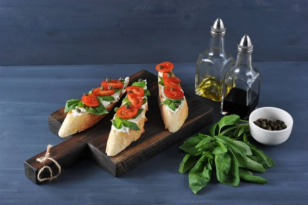 Bruschetta yumuşak peynir, taze domates, fesleğen yaprakları ve kapari ahşap bir tahta üzerinde. Çerçevede, zeytinyağı, balzamik sirke, fesleğen bir demet ve kapari ile bir fincan. Koyu arka plan. 
