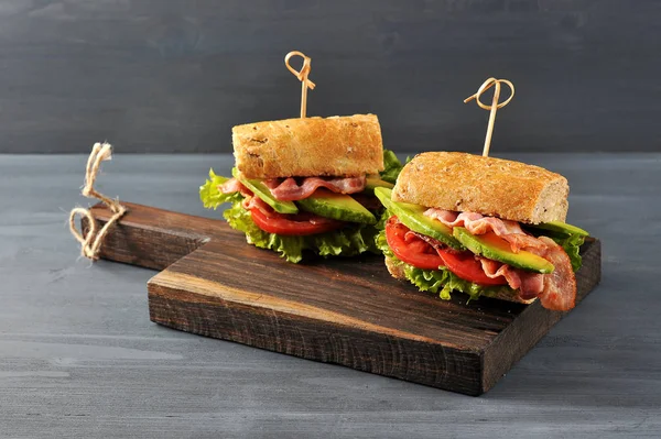 Appetizing sandwich on a wooden board. Baguette sandwich with filling from lettuce, slices of fried bacon, tomato and avocado. Dark wooden background. View from above. Close-up. Macro photography.