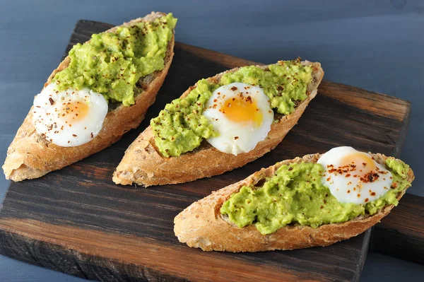 Appetizing bruschetta with egg and avocado on a wooden board. An exquisite alternative to a sandwich. On the fried baguette, the avocado pulp and fried quail egg. Dark background. Close-up.