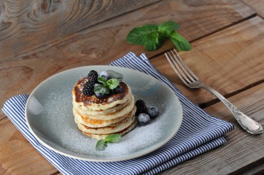 Iştah açıcı krep yaban mersini, böğürtlen, nane ile dekore edilmiş ve pudra şekeri serpilir. Tabağın altında bir peçete var, çatalın yanında. Ahşap arka plan. Close.
