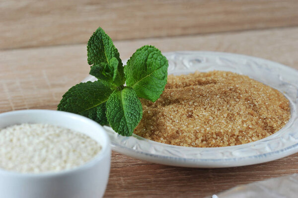 Тростниковый сахар на белой тарелке. Сахар украшен веточкой свежей мяты. Далее в белой чашке семена кунжута. Светлый фон. Крупный план. Макрофотография
.