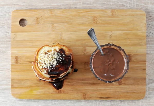 Pancakes are lined with a slide one on top of the other. Pancakes are poured with chocolate syrup and decorated with nut crumbs. Next to the bank with chocolate peanut butter. View from above. Closeup