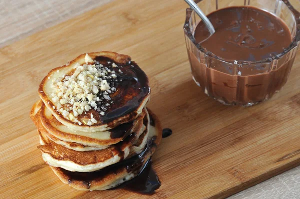 Pancakes are lined with a slide one on top of the other. Pancakes are poured with chocolate syrup and decorated with nut crumbs. Next to the bank with chocolate peanut butter. Closeup