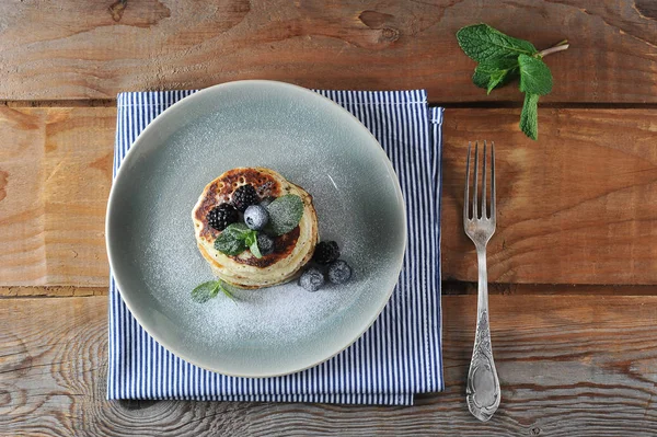 Iştah açıcı krep yaban mersini, böğürtlen, nane ile dekore edilmiş ve pudra şekeri serpilir. Tabağın altında bir peçete var, çatalın yanında. Ahşap arka plan. Yukarıdan görüntüle.