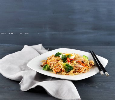 Düz bir tabakta brokoli ve tavuk eti ile Çin erişte. Erişte biber, mantar, havuç, yumurta içerir. Yemek çubuklarının çerçevesinde, peçete. Karanlık arka plan. Close.