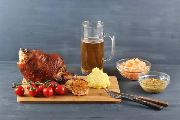 A traditional dish of Czech and German cuisine. Baked pork knuckle. In the frame a glass of beer, cherry tomatoes, mashed potatoes, grilled garlic, mustard, sauerkraut, cutlery. Dark background.